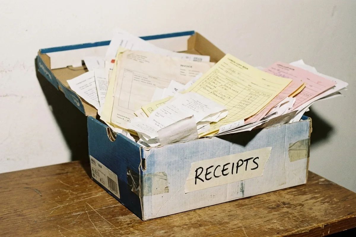 A worn shoebox labeled RECEIPTS, overflowing with crumpled paper invoices and receipt spools on a wooden desk.