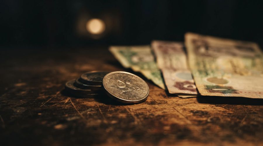 Qatari riyal notes and coins on a worn shop counter under a warm bulb, soft-focus in the dark.